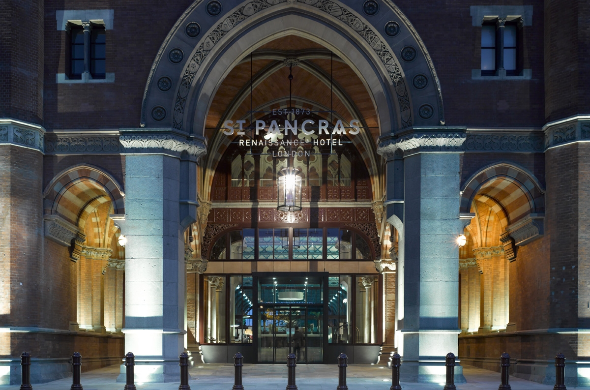 Illuminated entrance of St Pancras Renaissance Hotel in London. Gothic-style archway with ornate details, flanked by columns. Hotel name and establishment date visible above glass doors.