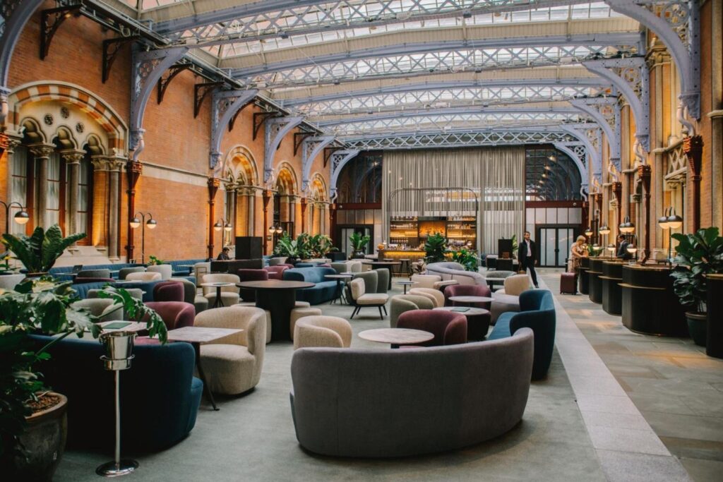 Elegant St Pancras Renaissance hall with vaulted glass ceiling, ornate iron arches, and brick walls. Comfortable seating area features modern curved chairs in muted colors. Plants and warm lighting create inviting atmosphere.