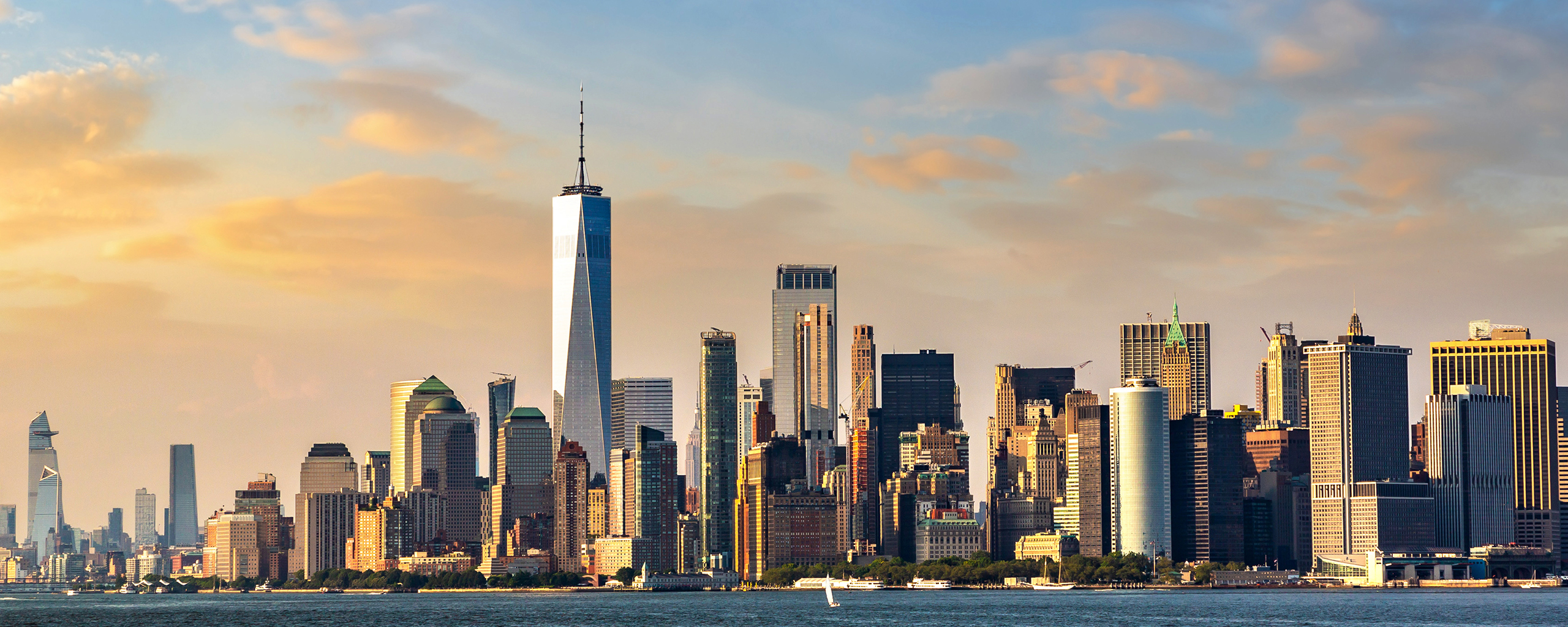 New York City skyline at sunset, featuring One World Trade Center and iconic skyscrapers. Golden light illuminates buildings against blue sky with wispy clouds over harbor waters.