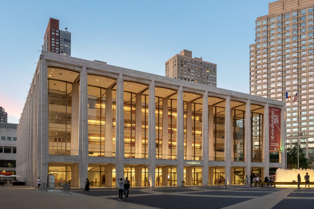 Lincoln Center's David Geffen Hall at dusk, illuminated facade with glass walls and columns, surrounded by New York City high-rises.